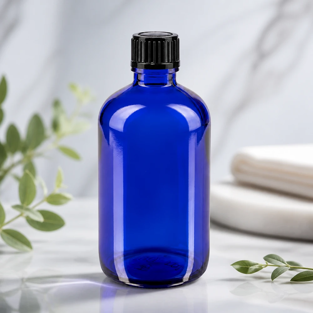 Apteekkipullo sininen 100ml tippapullo korkki 1mm musta standardi musta Sininen 100 ml:n lasipullo, jossa on uurrettu musta korkki, valkoisella marmoripinnalla ja eukalyptusoksilla