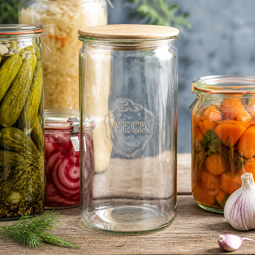 1590ml sylinteripurkki WECK RR100, jossa on puukansi WECK Zylinderglas 1590ml RR100 mit Holzdeckel zwischen Einmachgläsern mit Gurken Karotten und Sauerkraut auf Holztisch