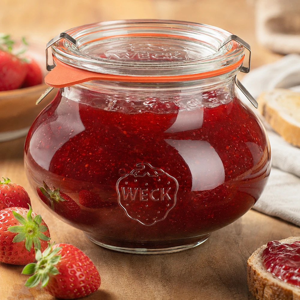 WECK koristeellinen purkki 560ml täynnä kotitekoista mansikkahilloa, kääntyvä kansi suljettu, ympärillä tuoreita hedelmiä ja vihanneksia.